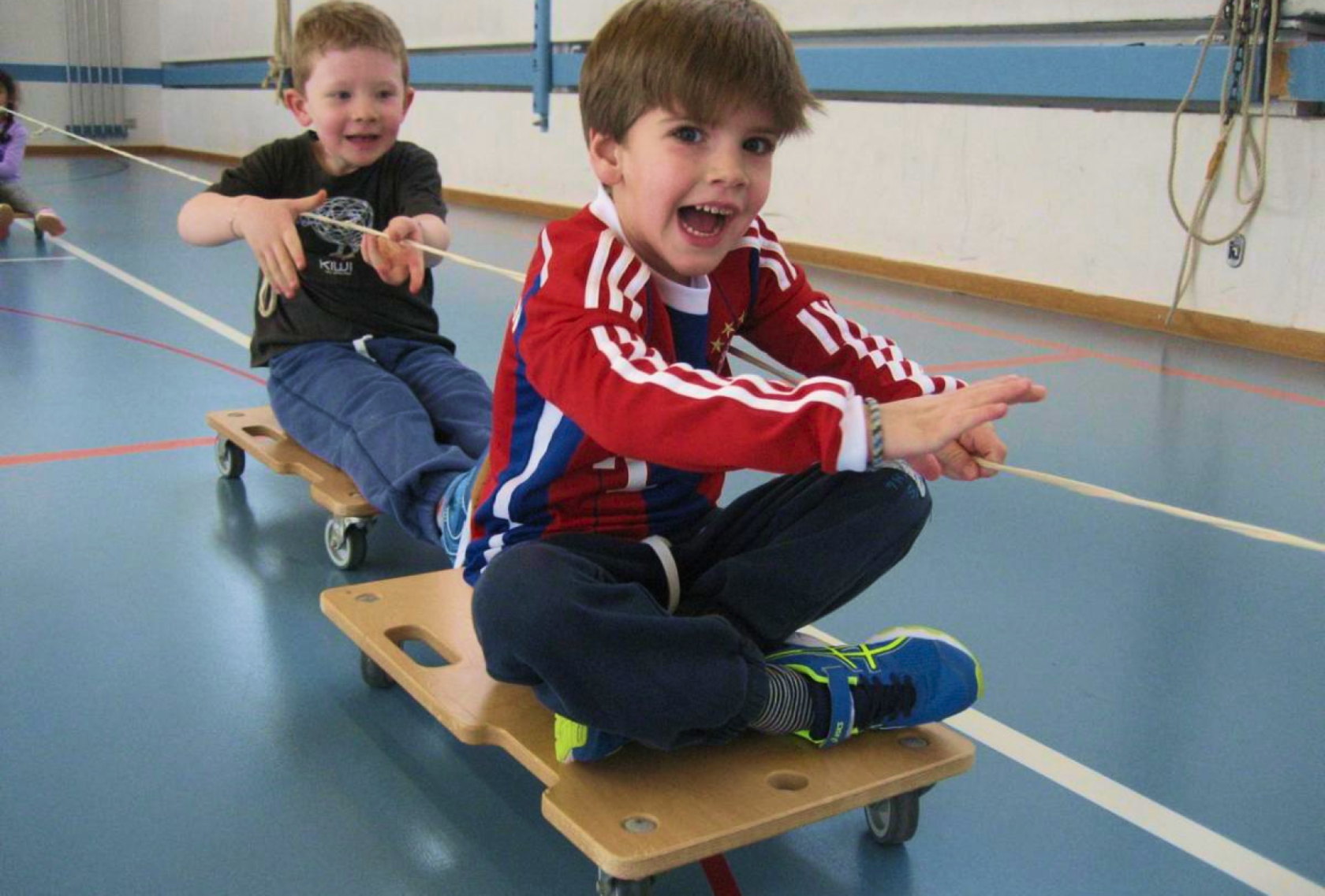 Zwei Jungen sitzen auf Rollbrettern in der Turnhalle und ziehen sich an einem Seil vorwärts